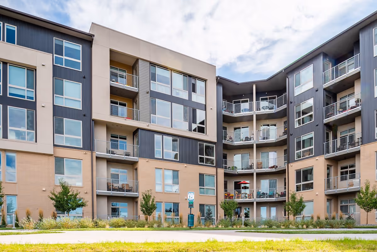 Exterior view of a modern multi-story residential building with balconies, large windows, and landscaped greenery in front under a partly cloudy sky.