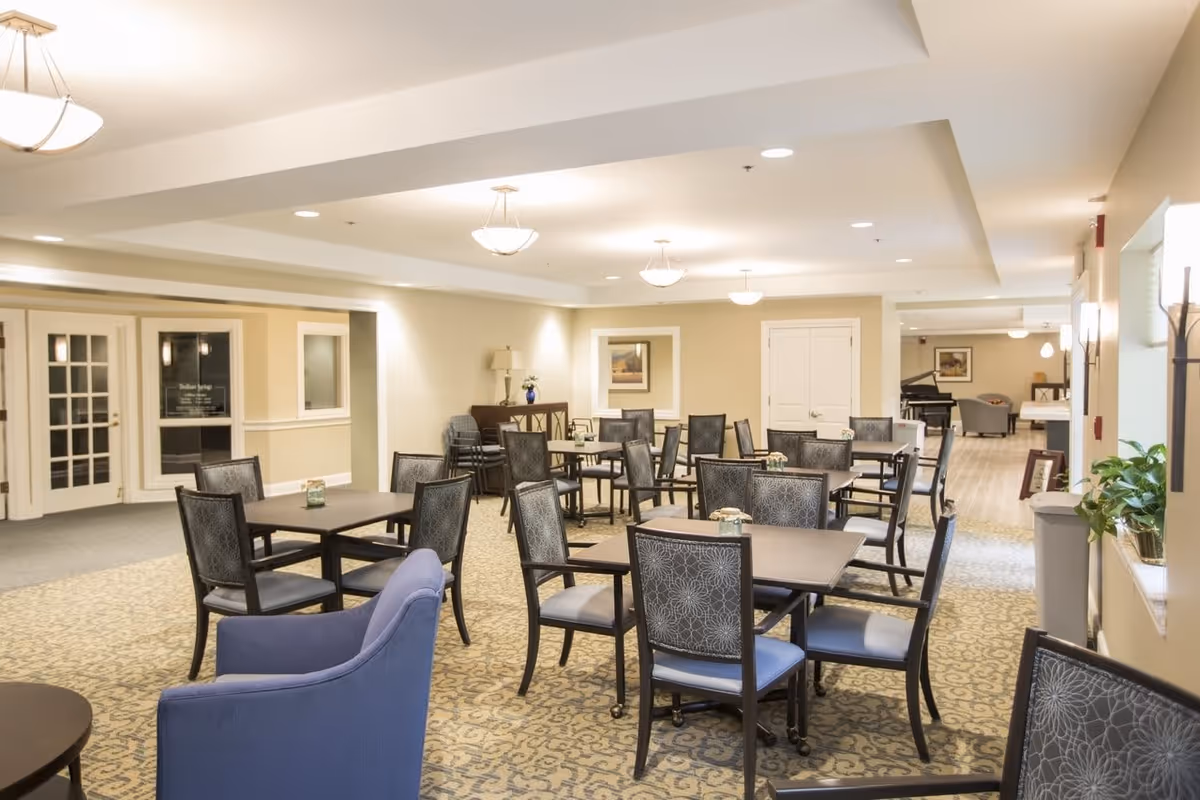 A well-lit dining area in a senior living facility with multiple square tables and cushioned chairs arranged neatly. The room has beige walls, patterned carpet, and ceiling lights. There is a blue armchair in the foreground and a piano visible in the background.
