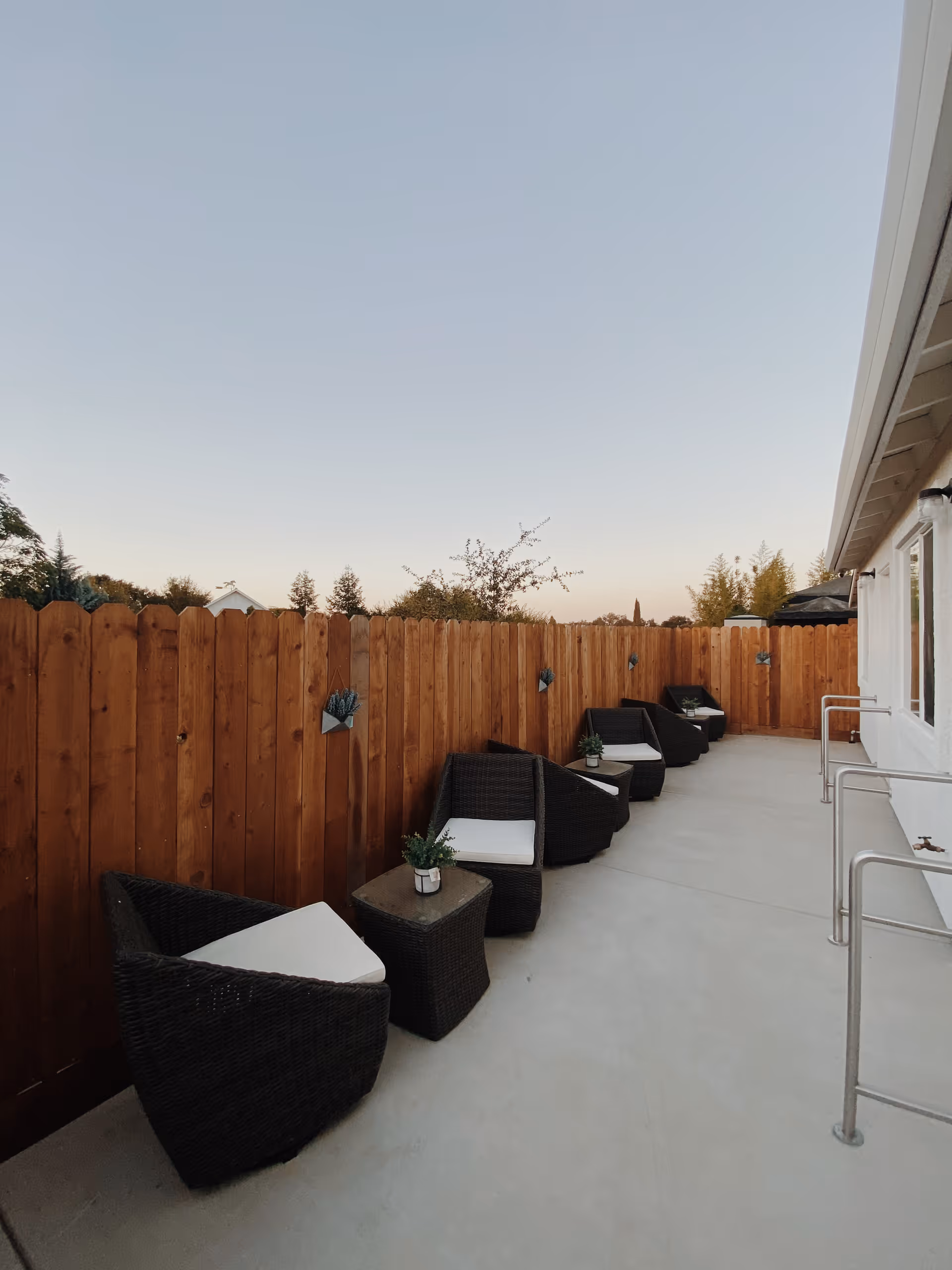 Outdoor patio area with a row of black wicker chairs with white cushions and small tables, each table holding a small potted plant. The patio is enclosed by a wooden fence and adjacent to a white building with metal handrails.