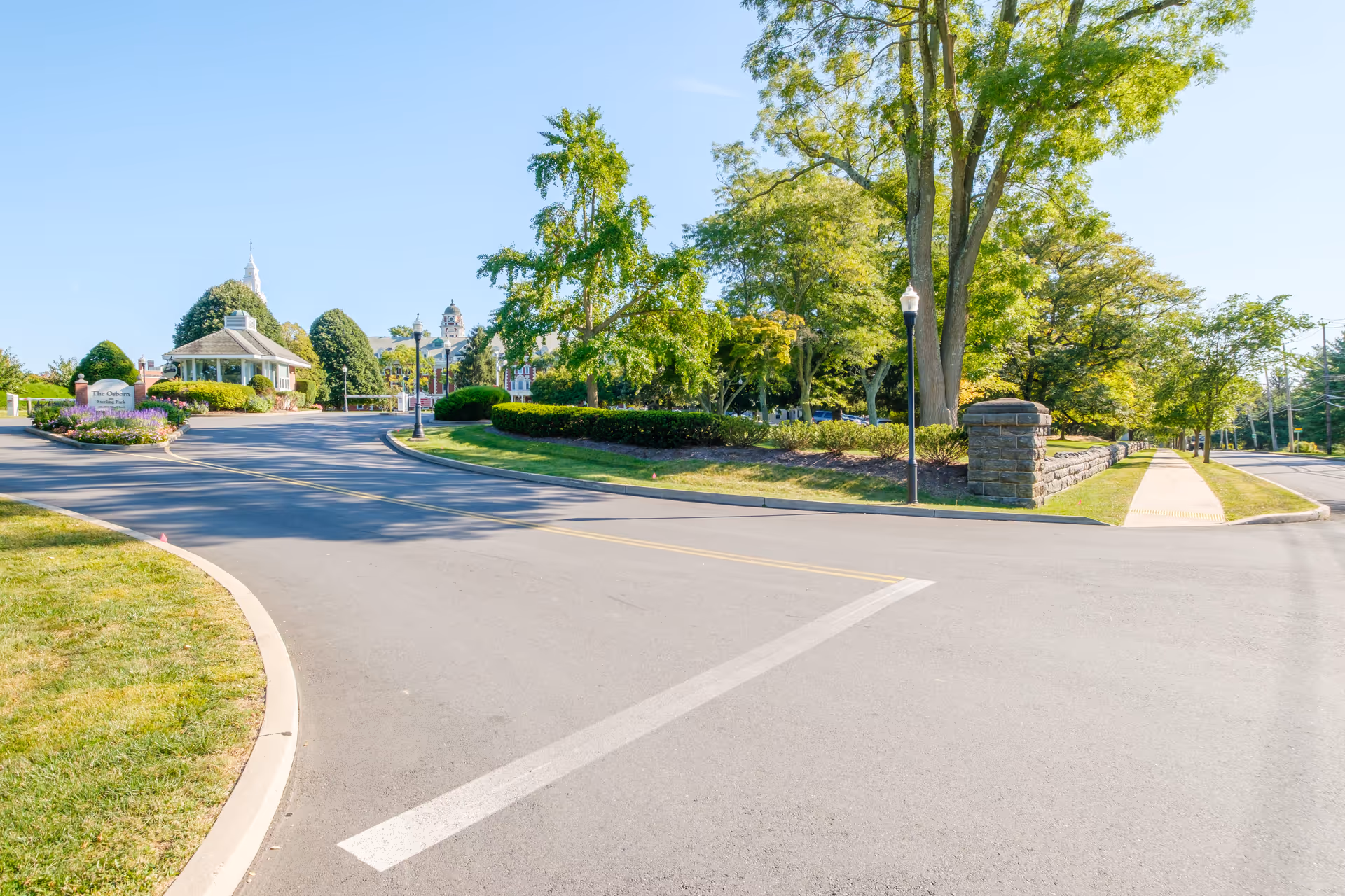 Landscaped, tree-lined driveway and entrance to a senior living community with a gazebo, stone sign, and sidewalk.