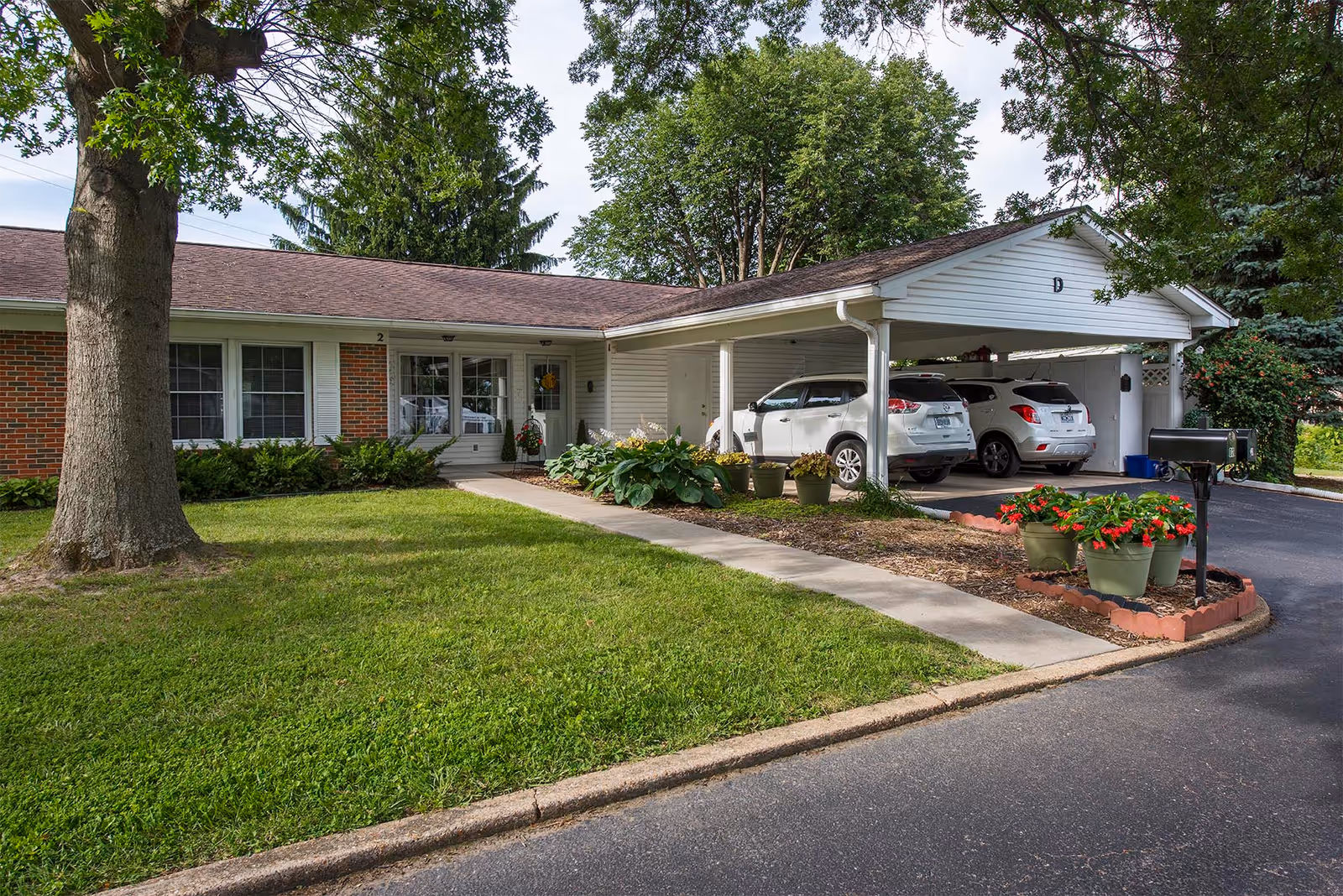 Front exterior of a single-story senior living building with a covered carport holding two white SUVs, a walkway and landscaped lawn.