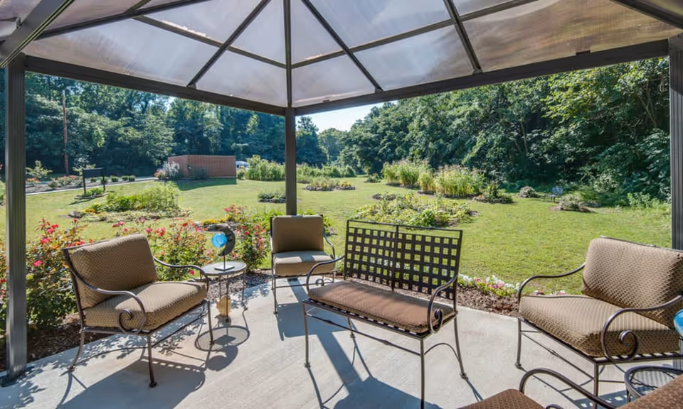Outdoor seating area under a transparent gazebo with cushioned metal chairs and a bench, overlooking a garden with green grass, flower beds, and trees in the background on a sunny day.