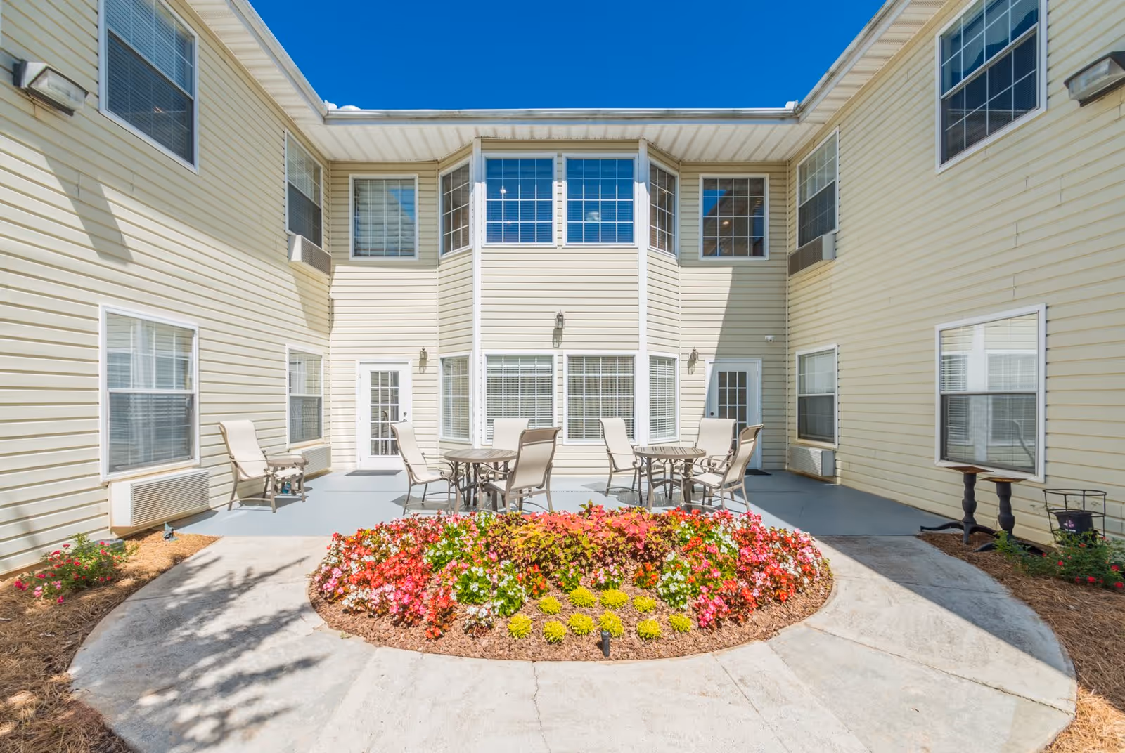 Outdoor courtyard area of a senior living facility with beige siding walls, multiple windows, and two doors. The courtyard features a circular flower bed with colorful flowers in the center, surrounded by a concrete walkway. There are several patio tables and chairs arranged around the flower bed for seating.