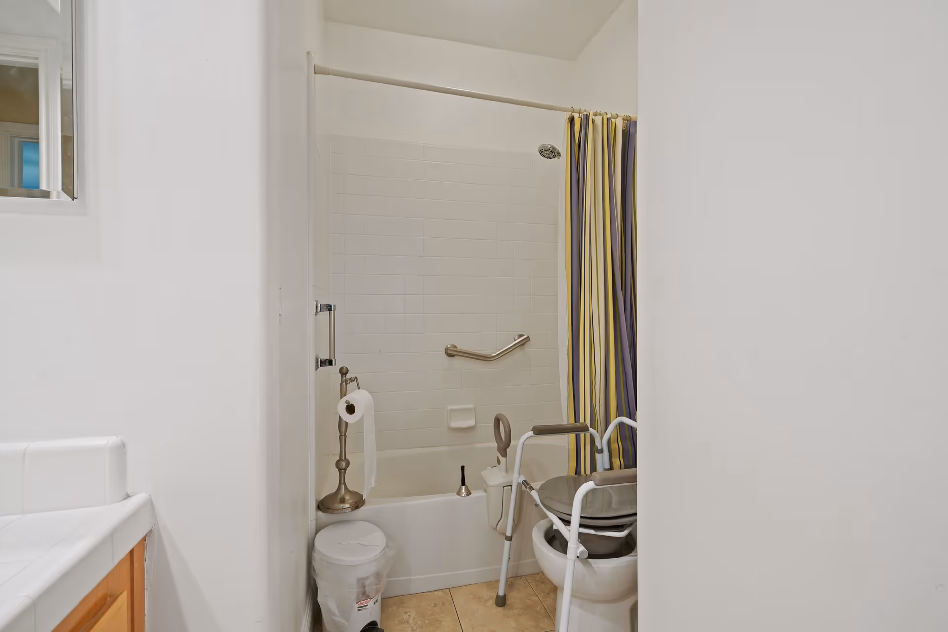 A small bathroom with a bathtub and shower combination featuring a striped shower curtain. There is a grab bar on the tiled wall inside the tub. A toilet with a raised seat and arm supports is positioned next to the tub. A toilet paper holder and a small trash can are also visible. The bathroom has white walls and a tiled floor.
