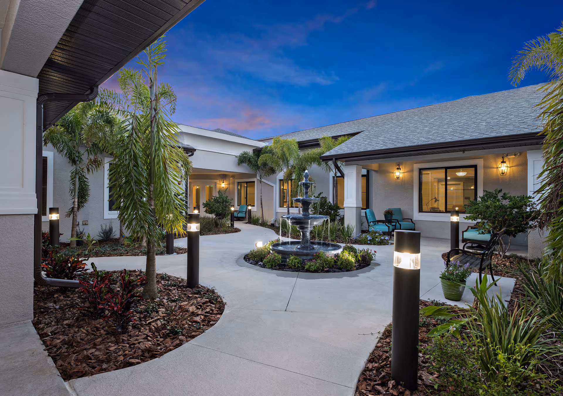 Outdoor courtyard area at dusk with a central three-tiered water fountain surrounded by landscaped plants and palm trees. The courtyard is bordered by a building with covered walkways, outdoor seating with turquoise cushions, and warm wall-mounted lights. Pathways with modern lamp posts lead through the garden beds.