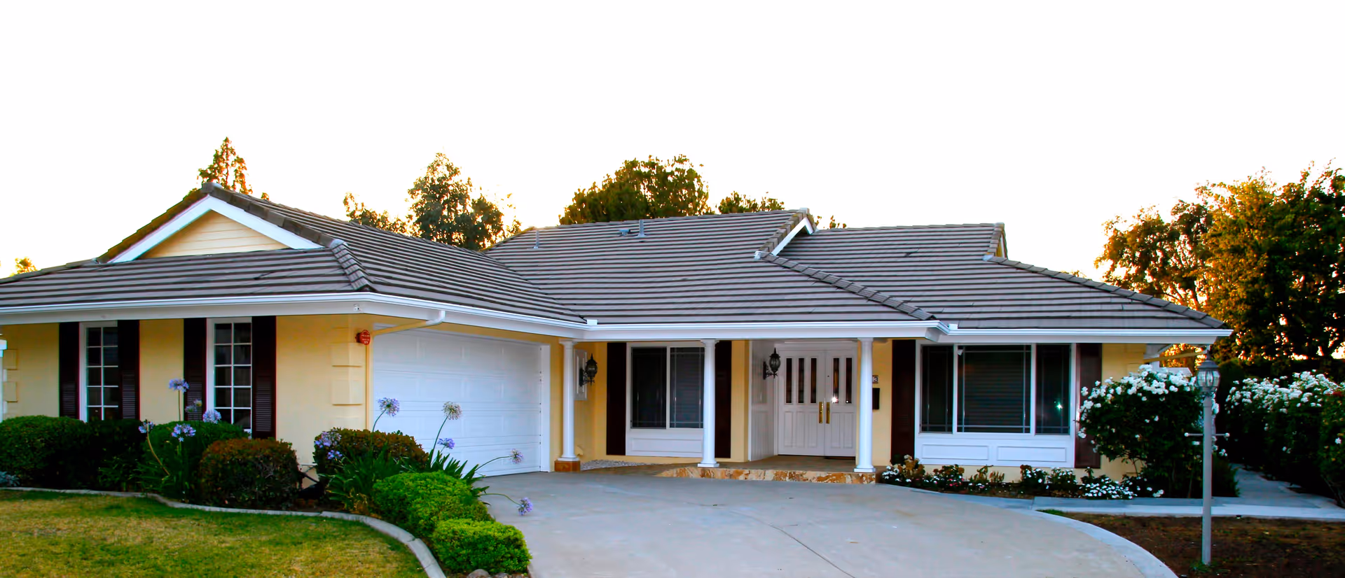 Front exterior view of a single-story residential building with a tiled roof, white double front doors, a two-car garage, and surrounding greenery including bushes and trees.