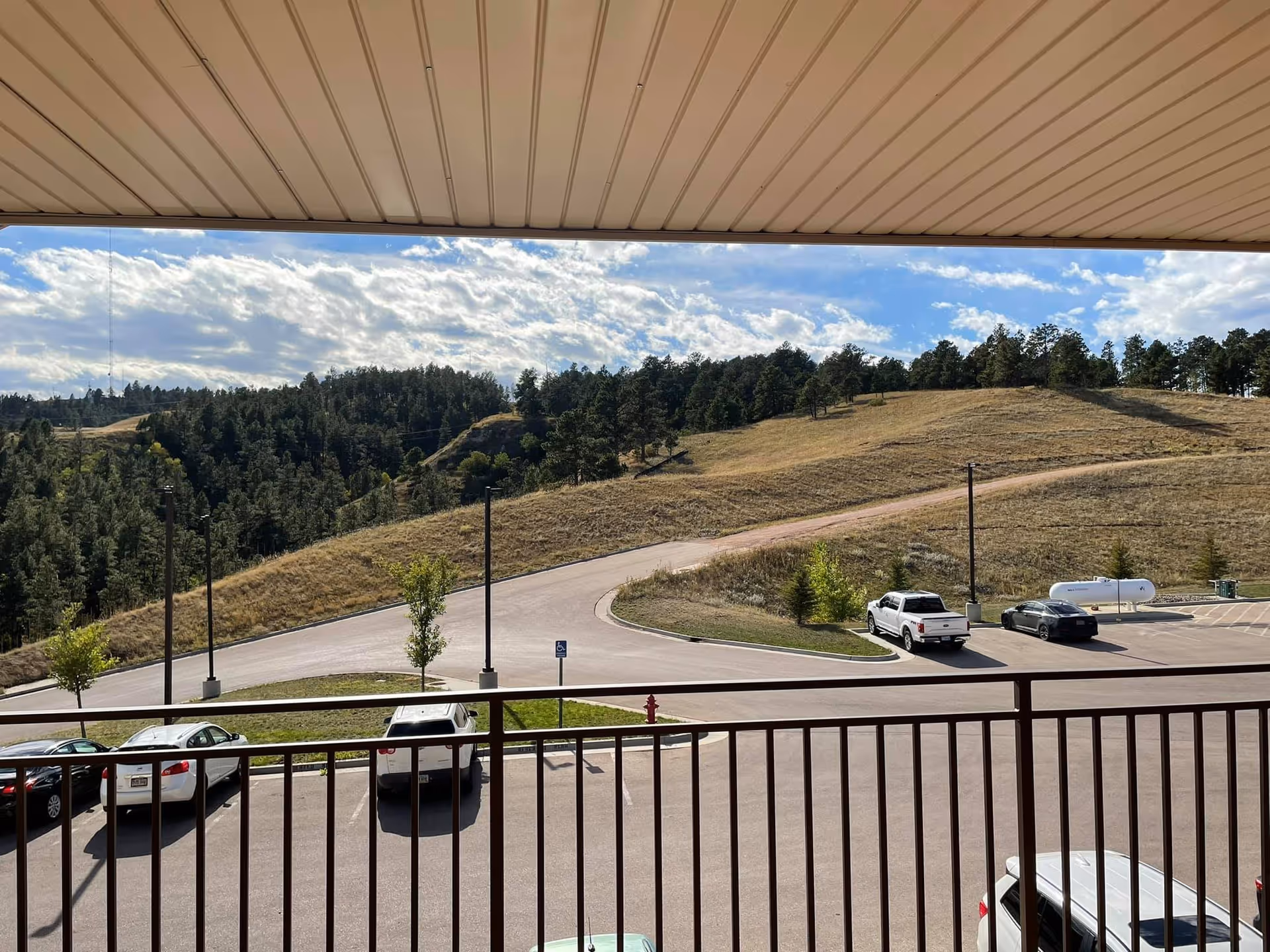 View from a covered balcony overlooking a parking lot with several parked cars and a scenic landscape of grassy hills and trees under a partly cloudy sky.