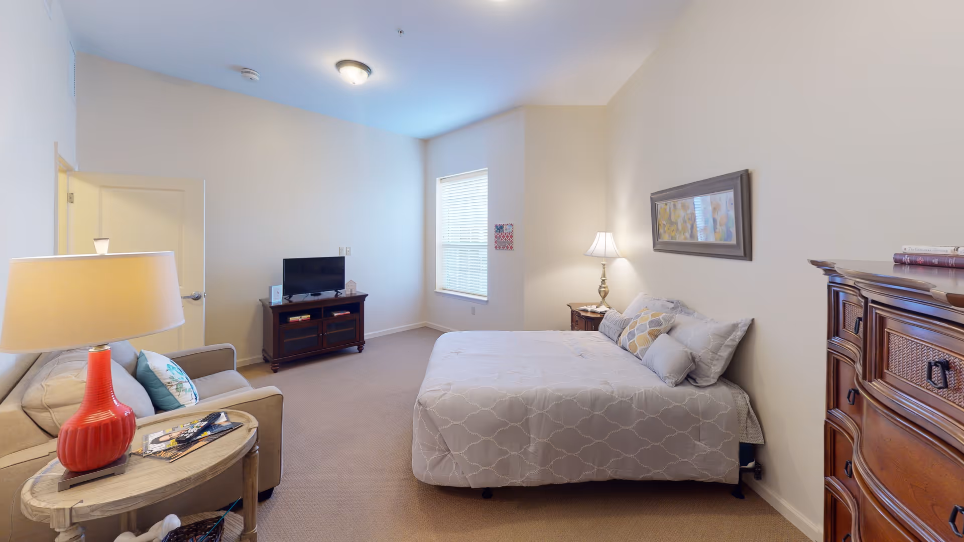 A cozy bedroom in a senior living facility featuring a bed with patterned bedding and multiple pillows, a wooden dresser, a nightstand with a lamp, a small TV on a wooden stand, a beige armchair with a side table holding a red lamp and magazines, and a window letting in natural light.