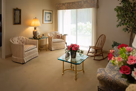 Cozy living room with two striped armchairs, a glass coffee table topped with flowers, a rocking chair, and sliding glass doors with vertical blinds.