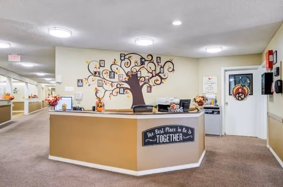 Reception area inside Winkler Court facility with a beige and brown front desk. The desk has a sign that reads 'The Best Place to Be Is TOGETHER'. Behind the desk is a wall decorated with a tree mural featuring framed photos. The hallway extends to the left with more decorations and lighting fixtures on the ceiling.