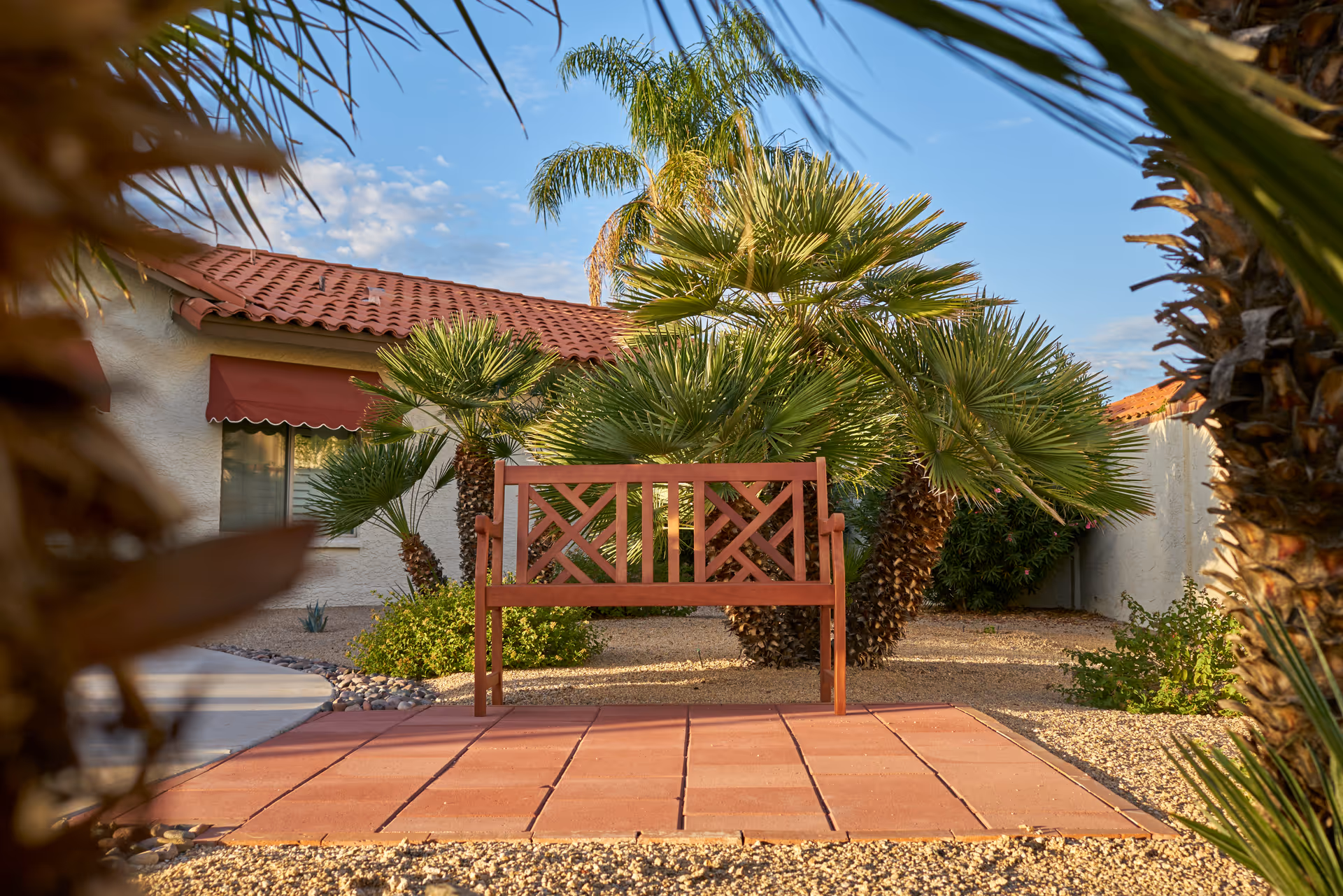 A wooden bench on a tiled patio area surrounded by palm trees and desert landscaping in front of a building with a red tiled roof and a window with a red awning.