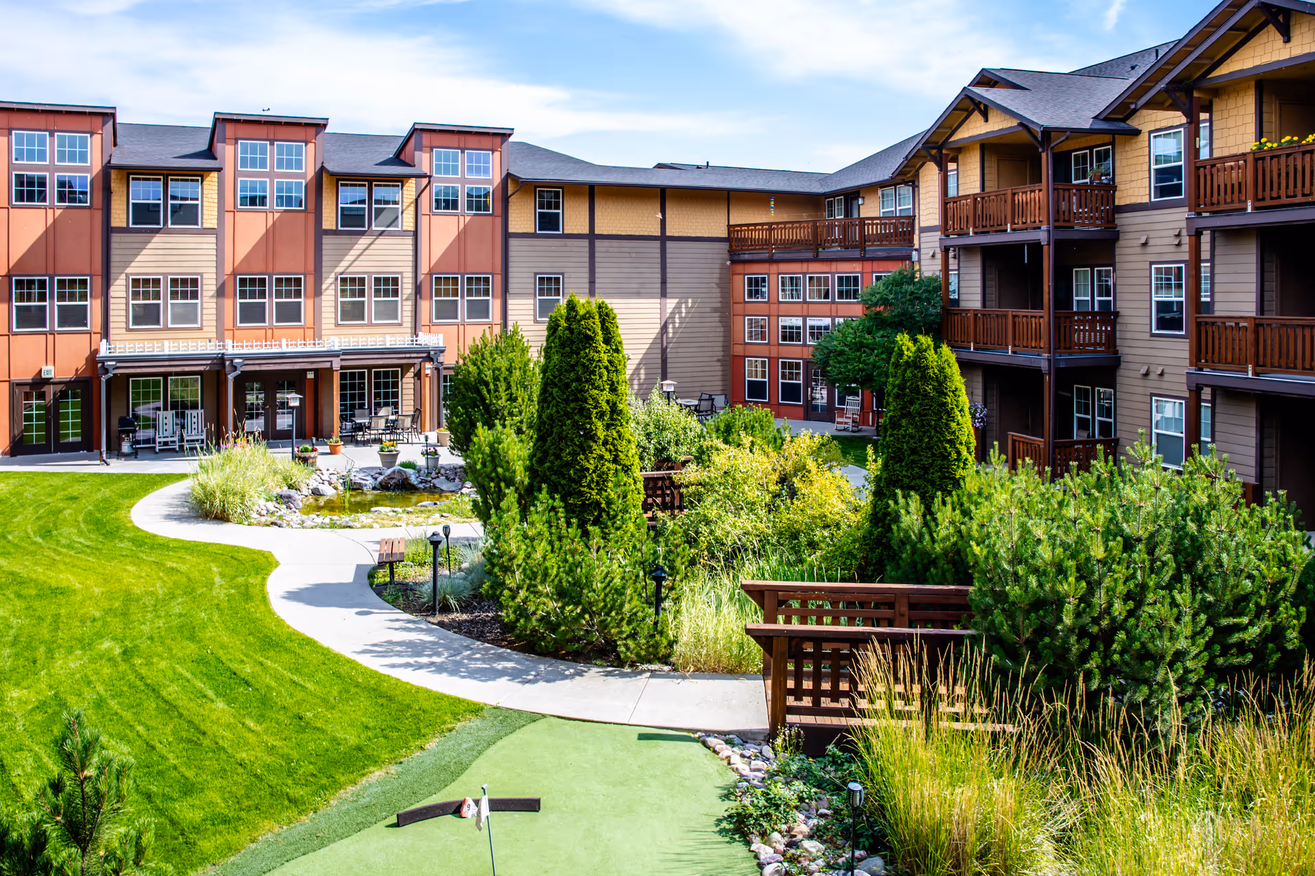 Outdoor courtyard area of The Springs at Missoula senior living facility featuring a well-maintained lawn, a small pond, walking paths, lush greenery, and a multi-story building with balconies and large windows surrounding the courtyard.