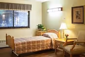 A single bedroom in a senior living facility with a bed covered in a brown patterned bedspread, a wooden nightstand with a lamp and a small plant, two upholstered chairs, a framed picture on the wall, and a window with blinds and a valance.