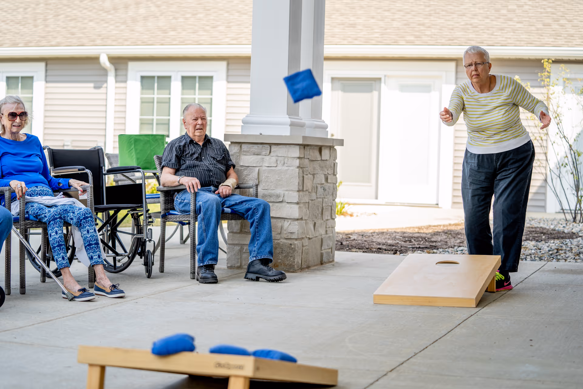 Senior residents seated under a covered patio watch a woman throw a bean bag toward a cornhole board.