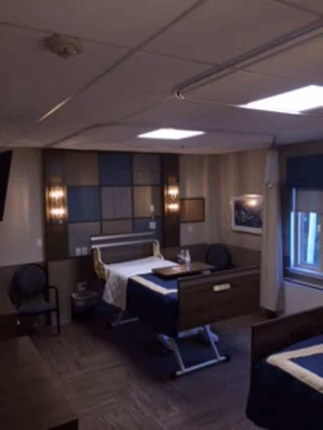 A dimly lit room with two hospital-style beds, each with a dark blue blanket and a white sheet. The room has a modern design with a patterned wall behind one bed, two wall-mounted lights, a window with a blue shade, and two black chairs. There is a tray table on one bed and a framed picture on the wall.