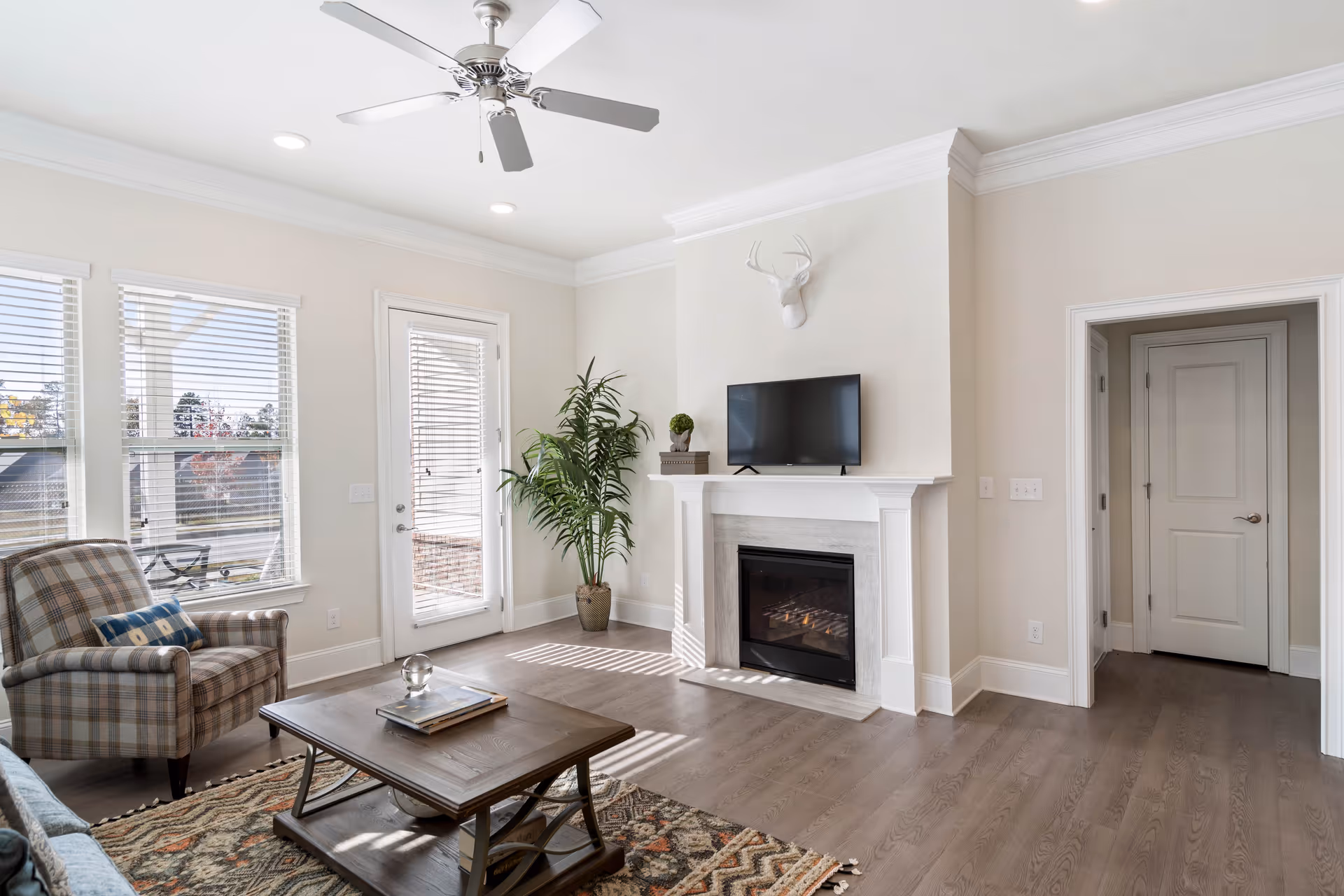 A bright living room with a ceiling fan, a plaid armchair with a blue pillow, a wooden coffee table on a patterned rug, a potted plant near a glass door, a fireplace with a mounted TV above it, and a white deer head decoration on the wall.