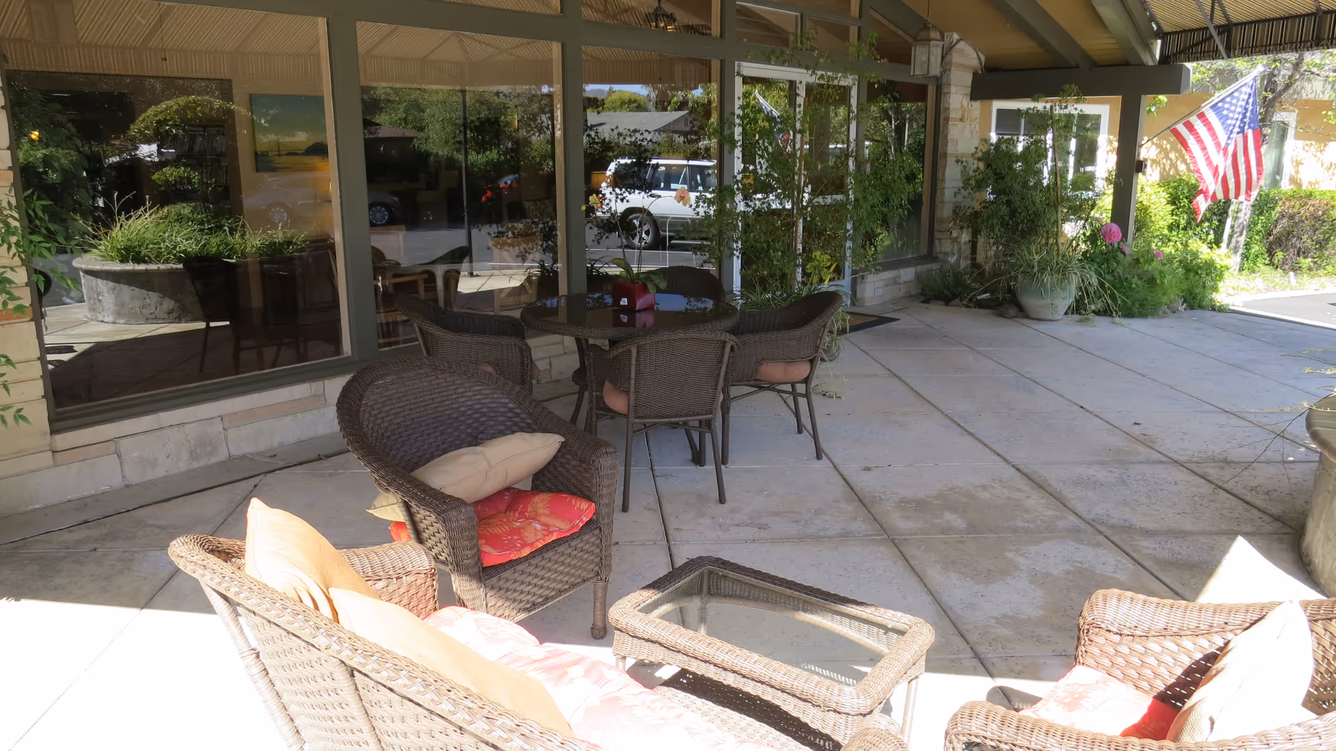 Outdoor covered patio area with wicker furniture including chairs and a glass-top table, potted plants, and an American flag near the entrance of a building with large windows reflecting the parking lot.