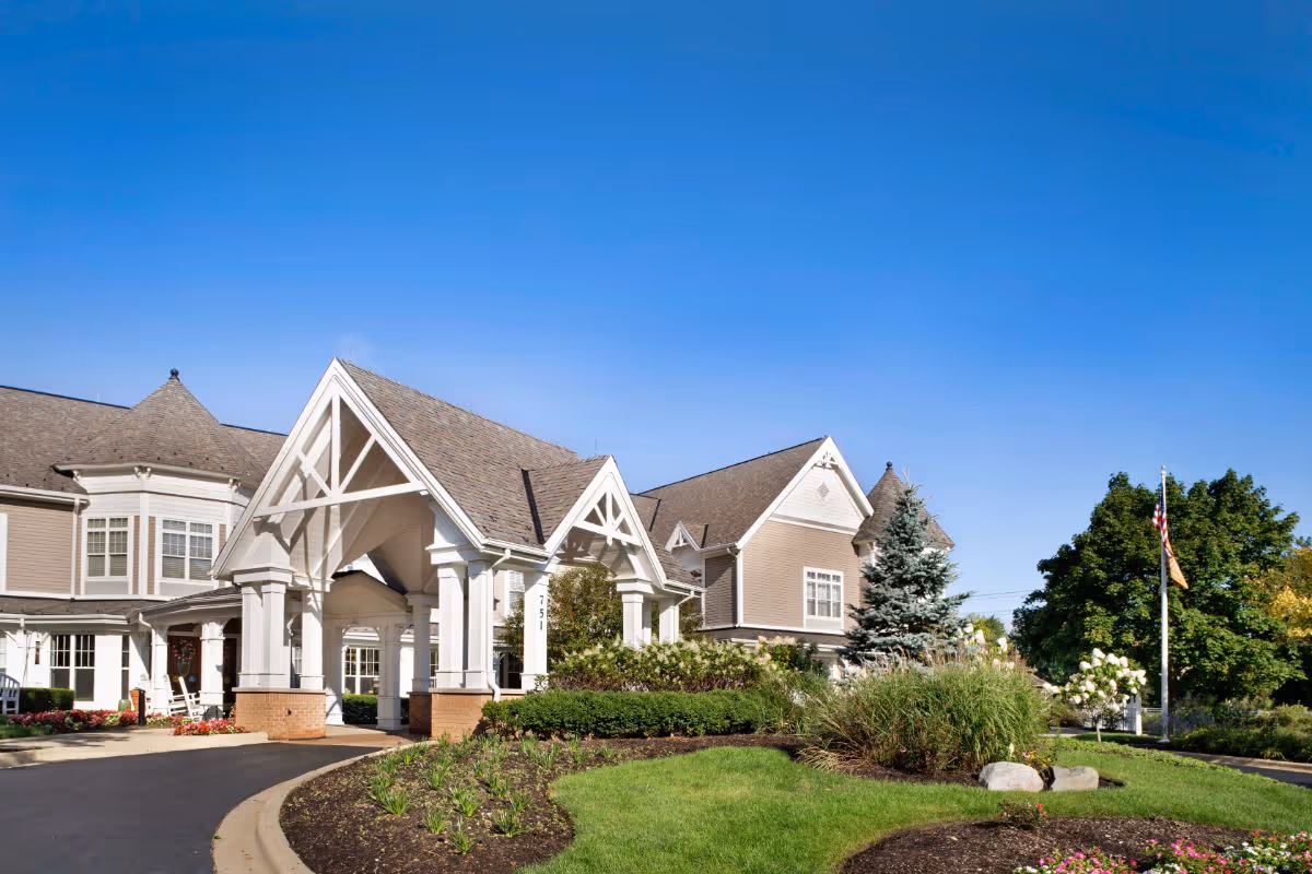 Front entrance and landscaped grounds of a senior living building with a covered porte-cochere under a clear blue sky.