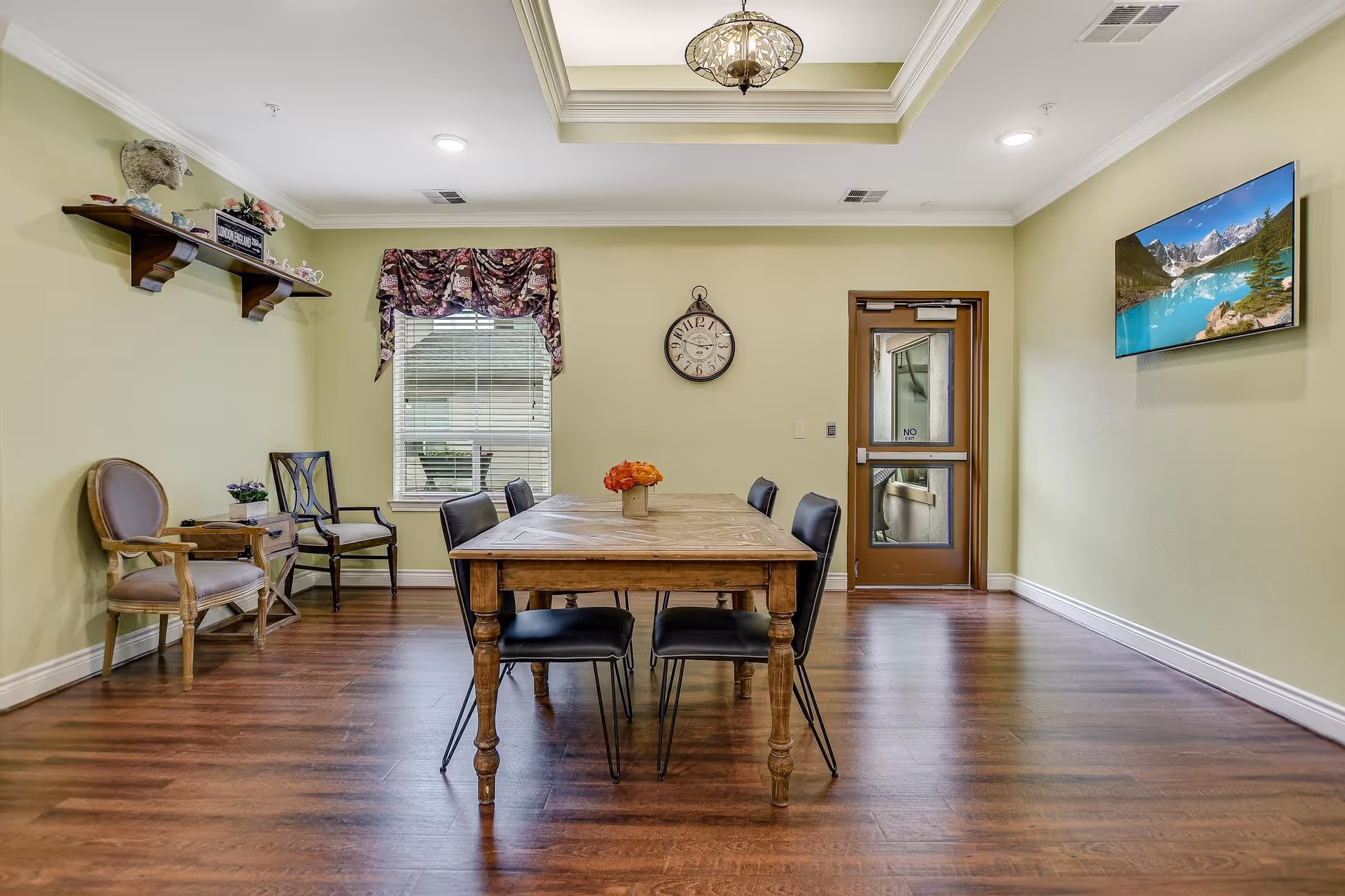 A small dining area with a wooden table and six black chairs, a window with floral valance, a wall clock, two additional chairs along the left wall, a shelf with decorative items, a door with a glass panel, and a wall-mounted TV displaying a scenic mountain and lake view. The room has light green walls and wood flooring.
