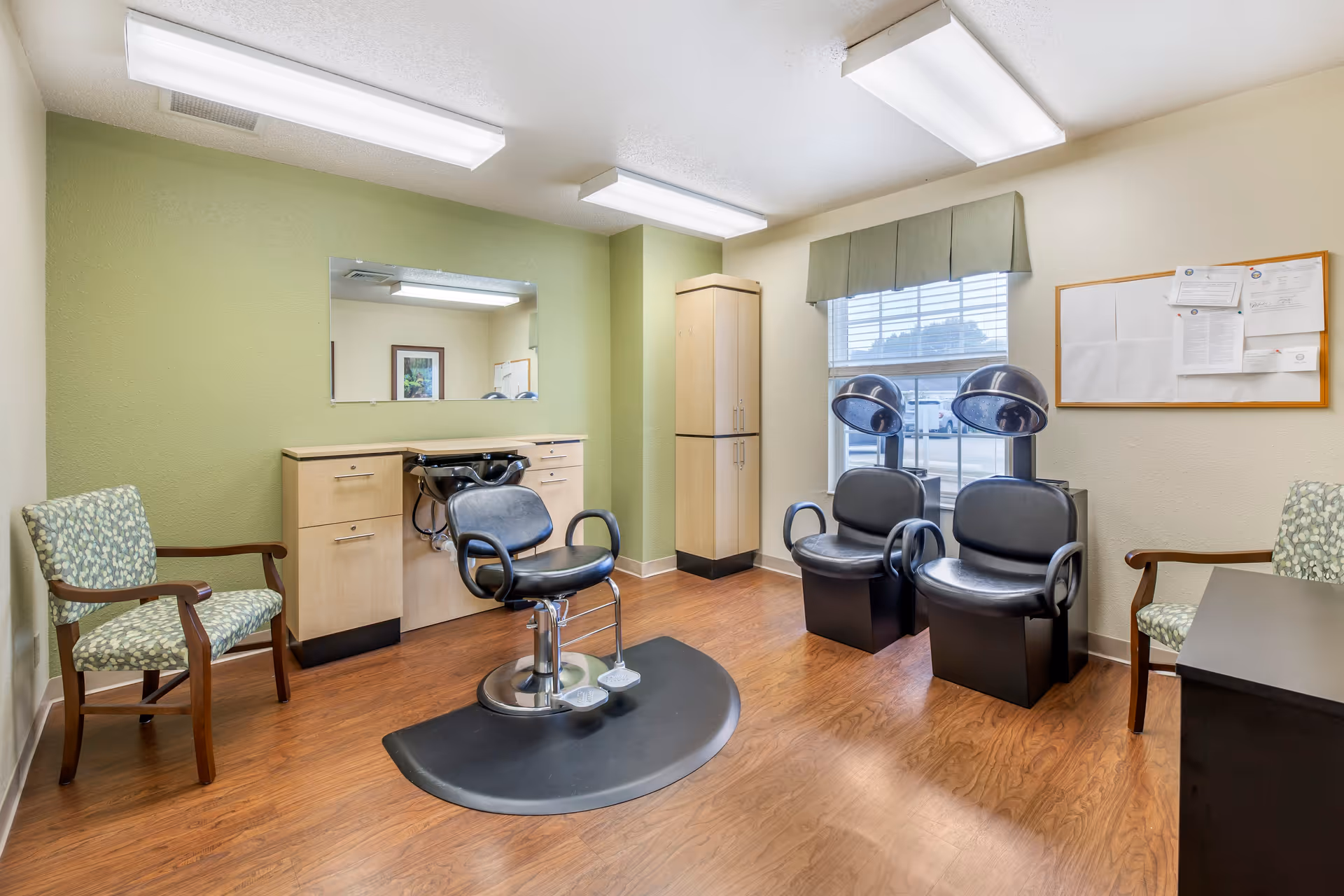 Interior view of a hair salon area in a senior living facility with a black salon chair in front of a sink and mirror, two black hair drying chairs near a window, two patterned armchairs, wooden flooring, and fluorescent ceiling lights.