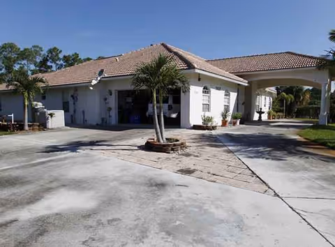 White single-story senior living building with a tiled roof, driveway and palm trees at the entrance.