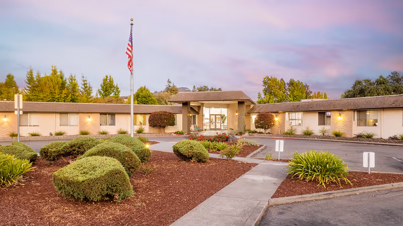 Exterior view of Santa Rosa Post-Acute facility at dusk with a circular driveway, landscaped bushes, an American flag on a flagpole, and a single-story building with lit windows and entrance.