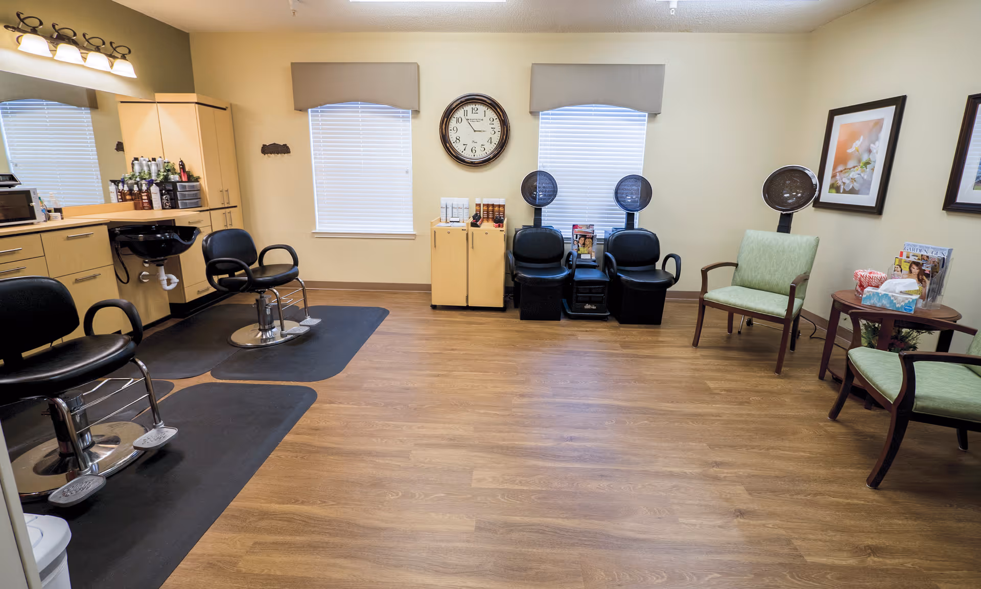 An interior hair salon area with styling chairs, hooded dryers, cabinets and a small waiting area with chairs and side tables.