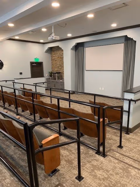 An auditorium-style room with several rows of wooden seats with black metal railings. At the front of the room is a large white projection screen framed by gray curtains. The walls are light-colored with dark trim, and there is a stone accent wall near a door with an illuminated green exit sign above it. A ceiling-mounted projector is visible above the seating area.