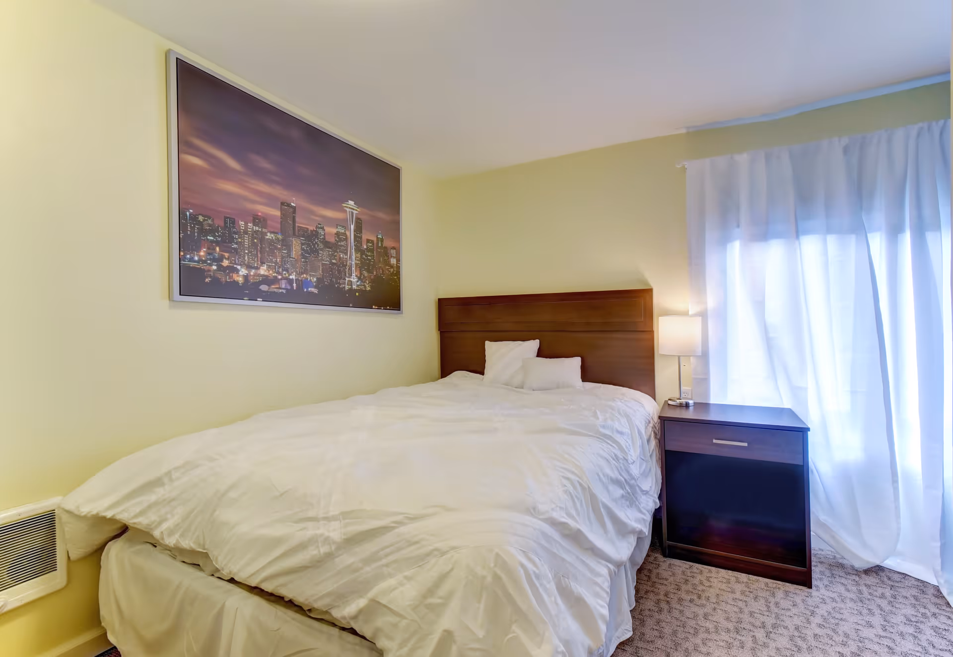 A bedroom with a neatly made bed featuring white bedding and two pillows. There is a wooden headboard and a dark wooden nightstand with a lamp on it. A large window with sheer white curtains allows natural light into the room. A framed picture of a city skyline at dusk hangs on the pale yellow wall above the bed.