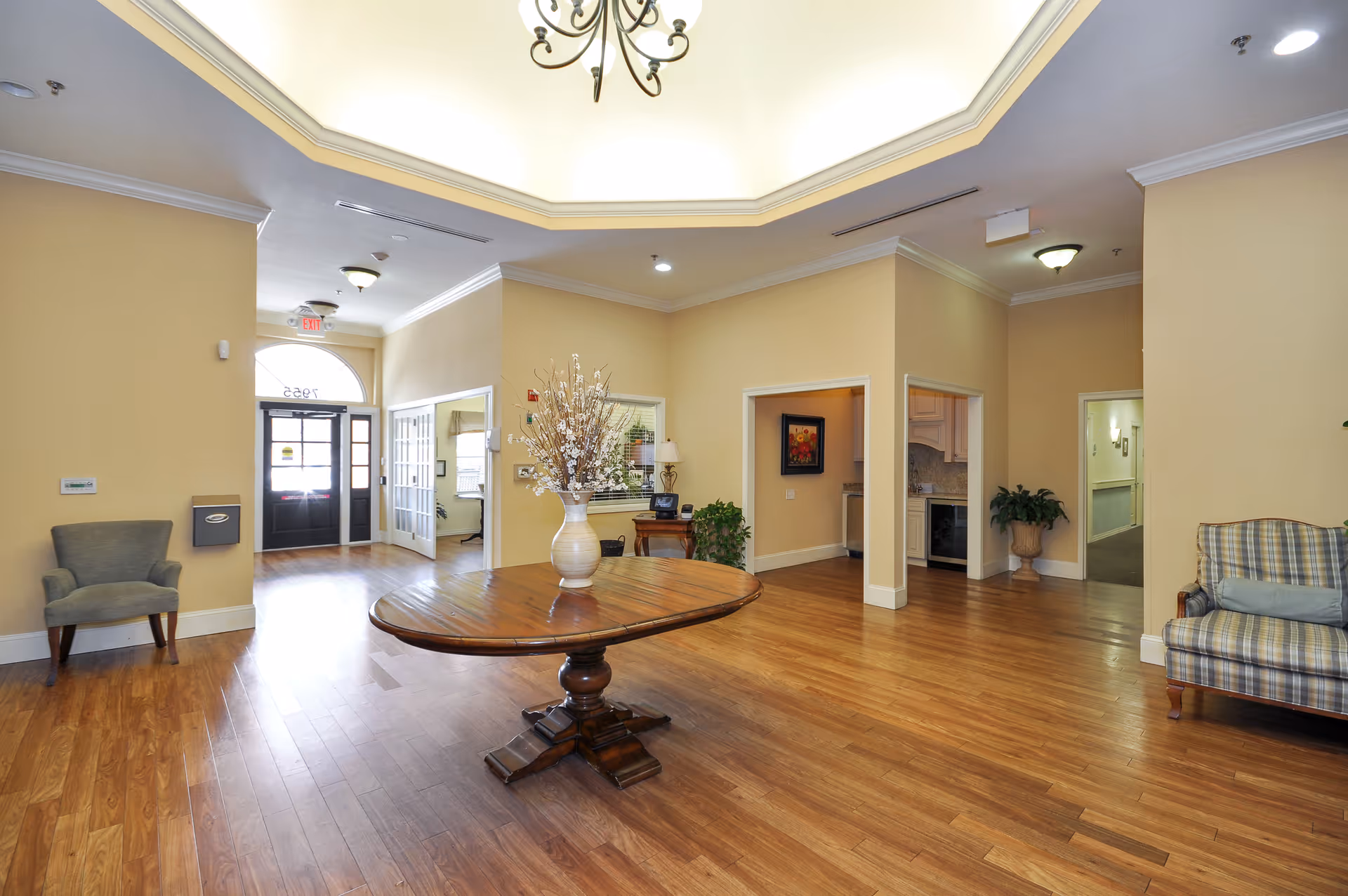 A spacious and well-lit interior common area with wooden flooring, beige walls, and a central wooden table with a large vase of flowers. There are two armchairs on either side of the room, a chandelier hanging from a recessed ceiling, and multiple doorways leading to other rooms. The entrance door with glass panels is visible in the background.
