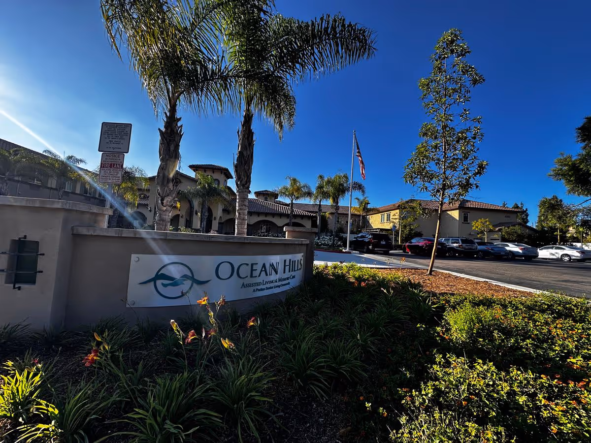 Front entrance signage and landscaped grounds of Ocean Hills Independent Living with palm trees, an American flag, and parked cars.