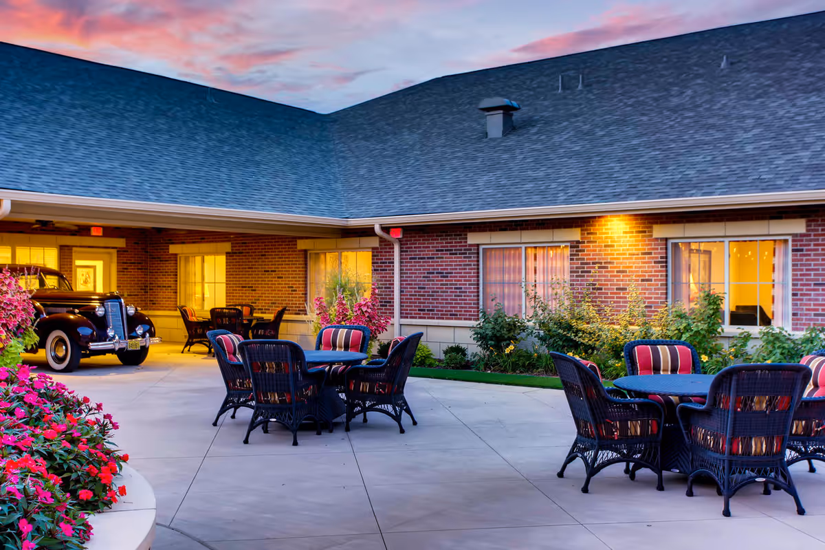 Outdoor patio area at Maple Glen Memory Care Community during sunset, featuring round tables with wicker chairs that have striped cushions, surrounded by flower beds and greenery, with a vintage car parked near the building's entrance.