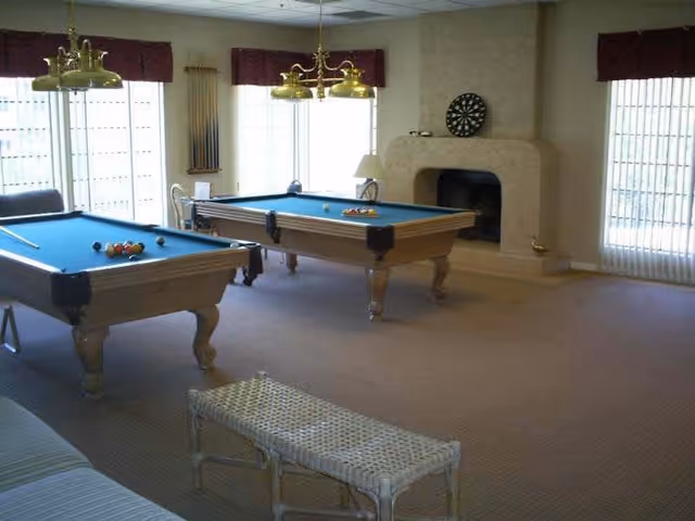 A recreational room with two pool tables, a dartboard mounted above a fireplace, large windows with vertical blinds, and a wicker bench in the foreground.