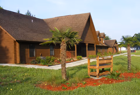 Front exterior of a brown wooden lodge with a walkway, landscaped beds, and small palm trees on a grassy lawn.