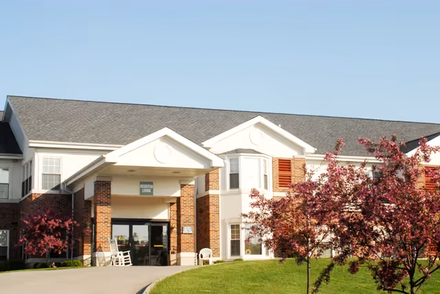 Exterior view of a two-story assisted living facility building with a covered entrance, brick and white siding facade, and blooming pink trees in the front lawn under a clear sky.