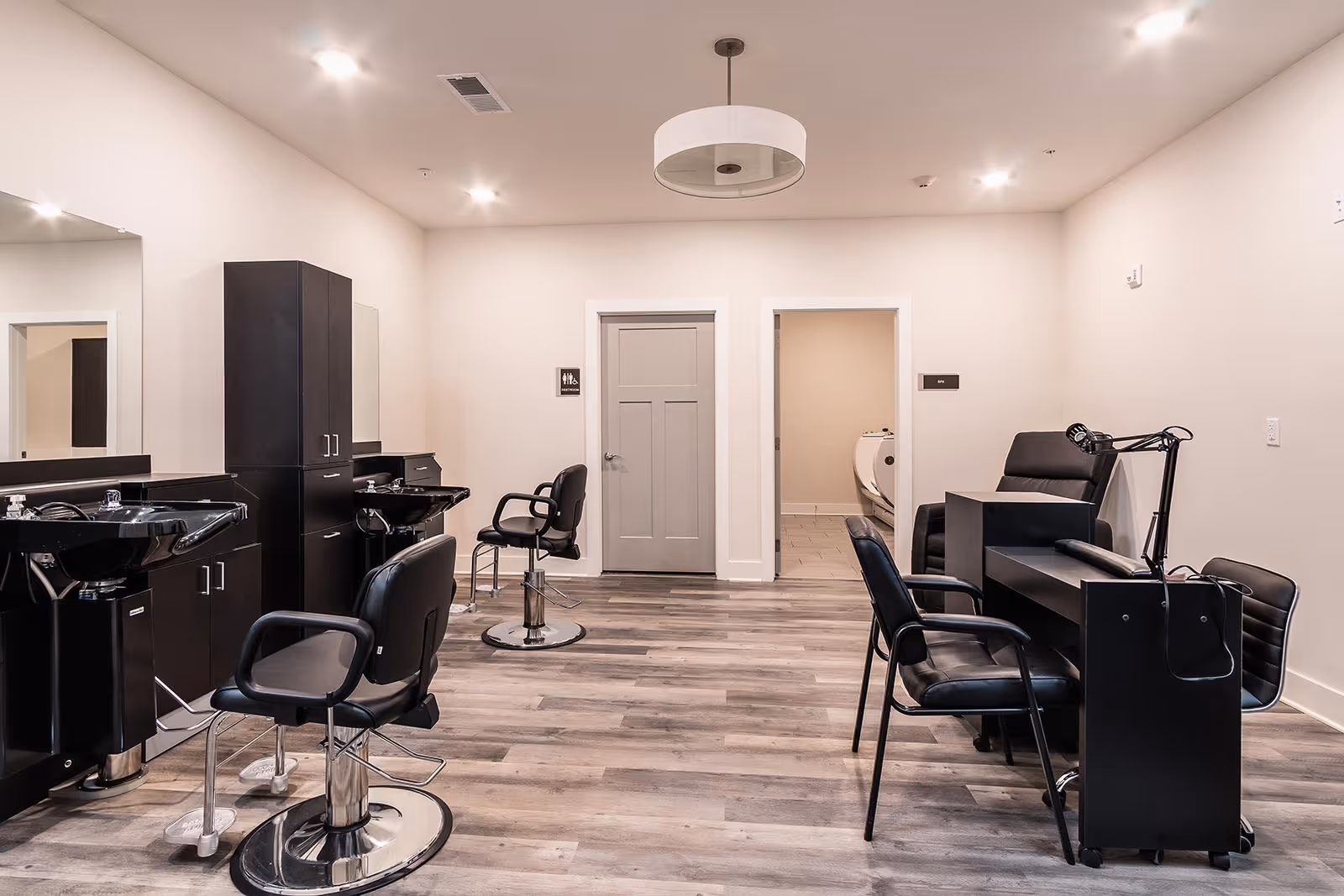 Interior view of a salon area in Canopy of Duval Station featuring black salon chairs, hair washing stations with sinks, a manicure table with chairs, and a comfortable recliner chair. The room has light-colored walls, wood flooring, and a modern ceiling light fixture. Two doors are visible at the back, one labeled restroom and the other labeled spa.
