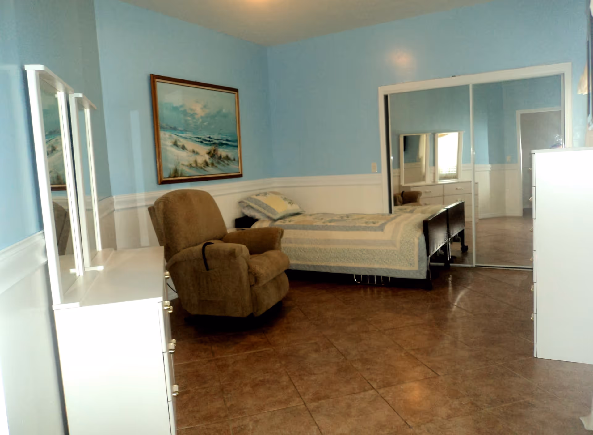 Light-blue bedroom with a recliner chair, a single bed, white dressers with mirrors, and mirrored closet doors.