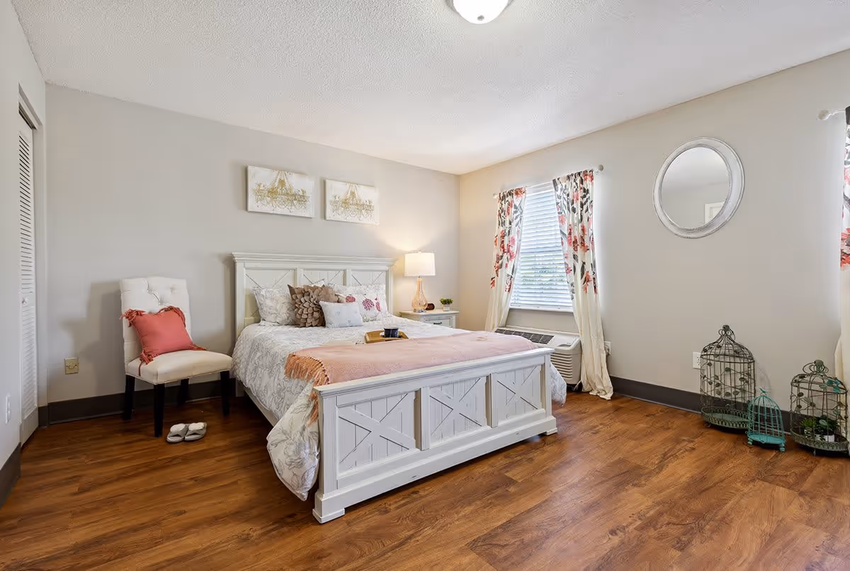 A bright and cozy bedroom with a white wooden bed frame, a bed with multiple pillows and a pink throw blanket. There is a white cushioned chair with a pink pillow, a nightstand with a lamp, floral curtains on the window, a round mirror on the wall, and decorative birdcages on the wooden floor.