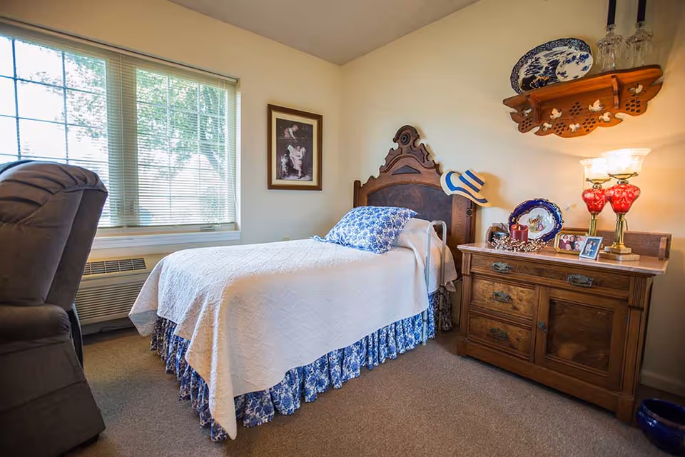 A cozy bedroom with a single bed featuring a wooden headboard, white quilt, and blue floral bed skirt and pillow. Next to the bed is a wooden nightstand with decorative items including a red lamp, framed photos, and a candle. A window with blinds lets in natural light, and a framed picture hangs on the wall. A cushioned chair is partially visible on the left side.