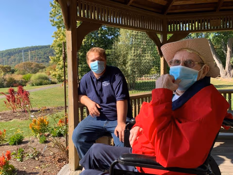 An elderly man wearing a red jacket, a straw hat, glasses, and a face mask sits in a wheelchair under a wooden gazebo. A younger man wearing a navy blue shirt and a face mask sits nearby on the gazebo's bench. Behind them is a garden with flowers and greenery, with hills visible in the background under a clear blue sky.