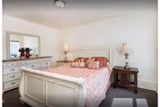 A bedroom with a white wooden bed frame and a pink patterned bedspread. There are multiple pillows on the bed. To the left of the bed is a white dresser with a large mirror above it and a vase with red flowers. On the right side of the bed is a wooden nightstand with a lamp. The walls are white and the floor is carpeted in a dark color.