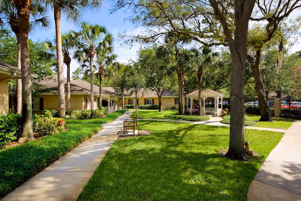 Sunny landscaped courtyard with walkways, benches, palm trees and a white gazebo in front of single-story buildings.