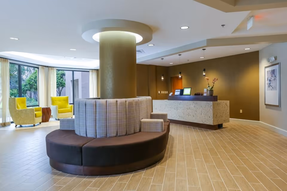 A modern senior living facility lobby with a circular seating area around a large column in the center. The seating has plaid cushions and dark brown base. In the background, there is a reception desk with three pendant lights hanging above it, a vase with flowers, and two computer monitors. To the left, there are large windows with sheer curtains and two yellow armchairs with a small wooden table between them. The floor is covered with light brown tiles arranged in a staggered pattern.