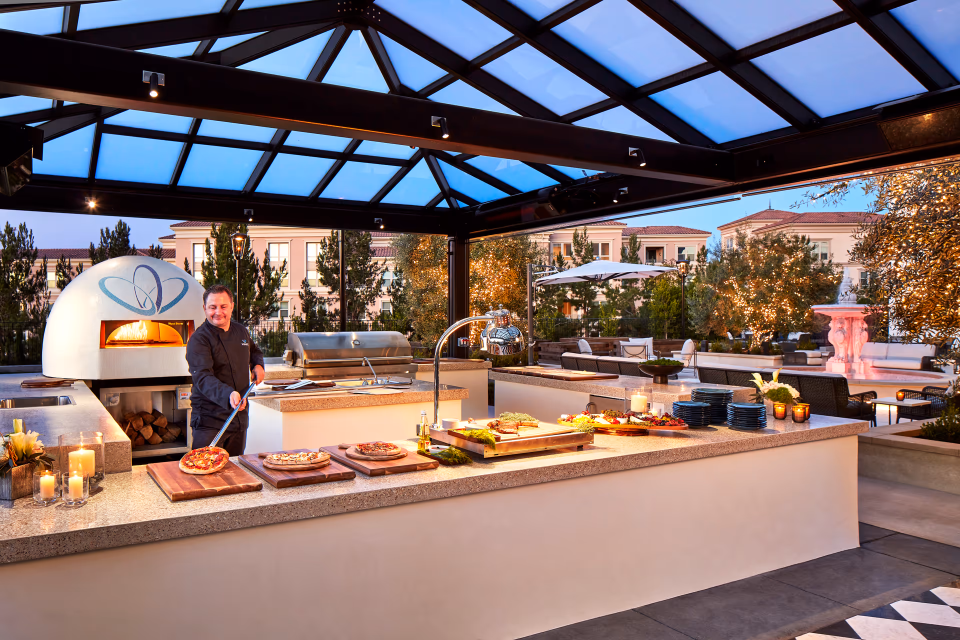 An outdoor covered kitchen area at Vivante Newport Center with a chef preparing pizzas near a wood-fired oven. The countertop displays several pizzas, plates, candles, and food platters. In the background, there are trees decorated with lights, outdoor seating, and a fountain, with residential buildings visible beyond.