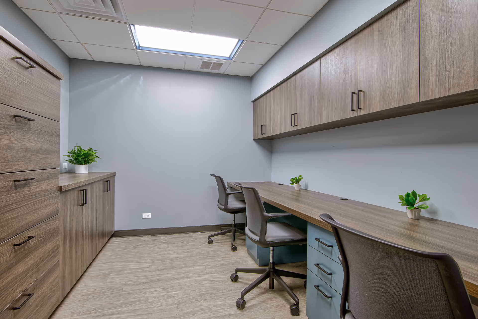A modern office or workroom with a long wooden desk, three office chairs on wheels, overhead wooden cabinets, and additional storage cabinets along the left wall. The room has light gray walls, a light wood floor, and a ceiling light panel.