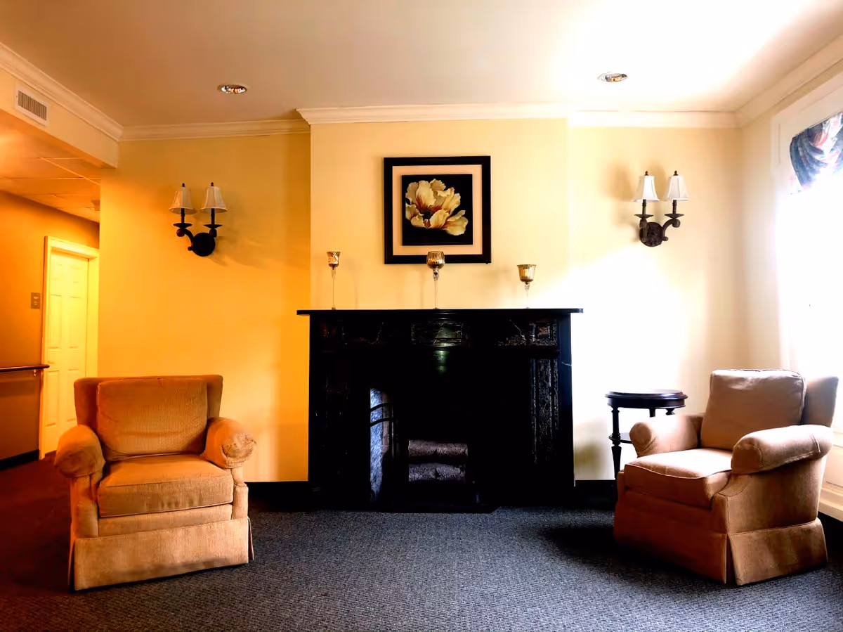 A cozy interior room with two beige upholstered armchairs facing each other on either side of a black fireplace. Above the fireplace is a framed floral artwork and three decorative candle holders. The walls are painted light yellow, with two wall-mounted lamps on either side of the fireplace. A small round side table is positioned next to the right armchair, and a window with a valance allows natural light to brighten the room.