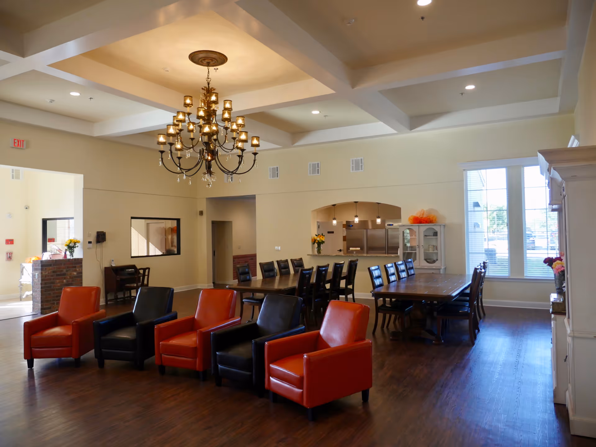 Spacious senior living common area with red and black armchairs, long dining tables, a chandelier, and large windows.