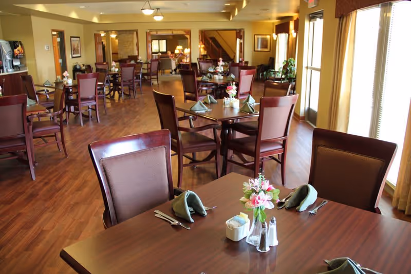 A spacious dining room with multiple wooden tables and chairs arranged neatly. Each table is set with green folded napkins, silverware, salt and pepper shakers, and a small vase with pink flowers. Large windows on the right side allow natural light to fill the room, and the floor is covered with wood-style flooring. The room has warm lighting and a cozy ambiance.