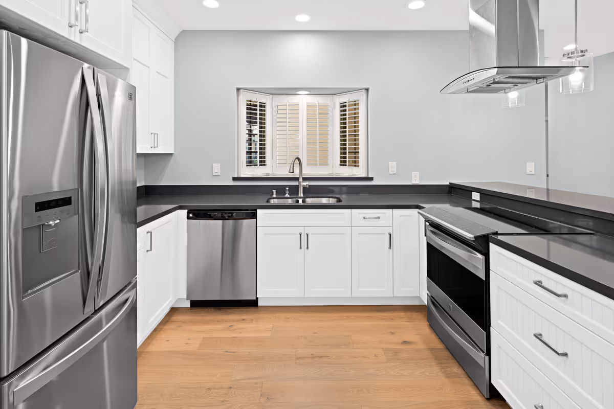 Modern kitchen with stainless steel refrigerator, dishwasher, and oven. White cabinets with silver handles, black countertops, a window with white shutters above the sink, and wooden flooring.