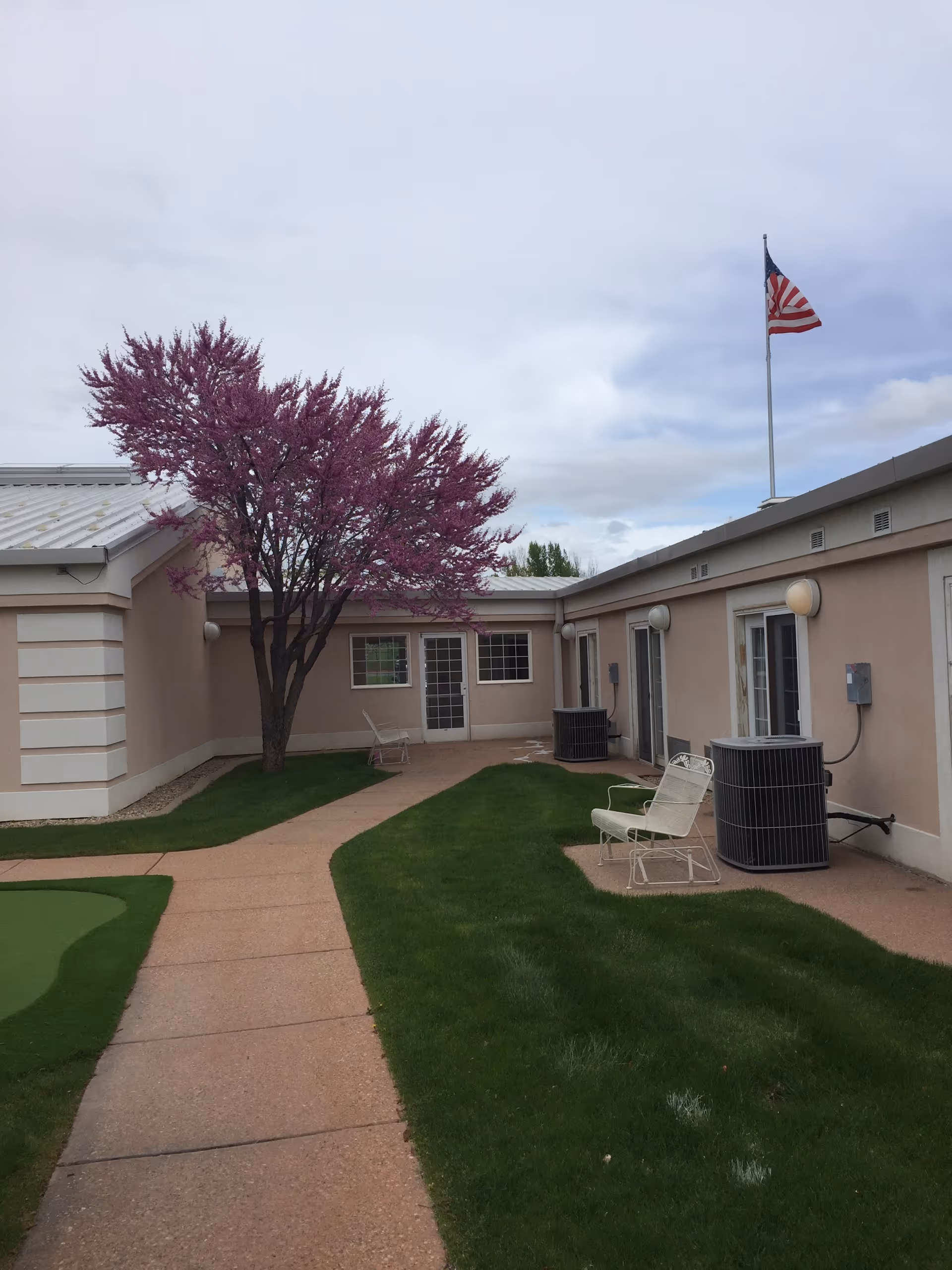 Outdoor courtyard of a senior living facility with a pink-blossomed tree, paved walkway, patio chairs, air conditioning units, and an American flag on a pole.